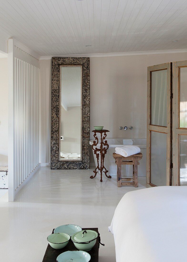 Vintage Schüsseln am Bettende auf Truhe und raumhoher Spiegel mit Rahmen an Wand lehnend neben Badewanne im Badbereich