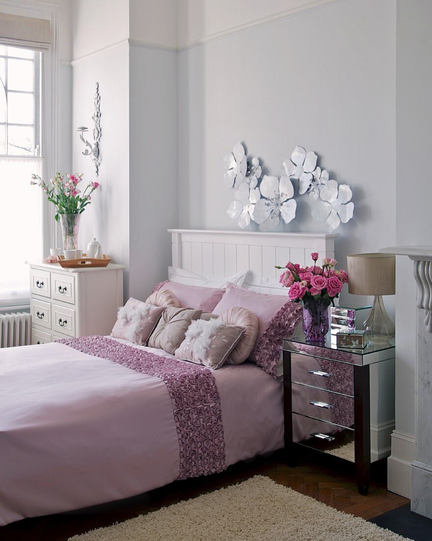 Doppelbett mit zartrosa Decke, silberner Nachttisch und weiße Kommode in einem elegantem Schlafzimmer