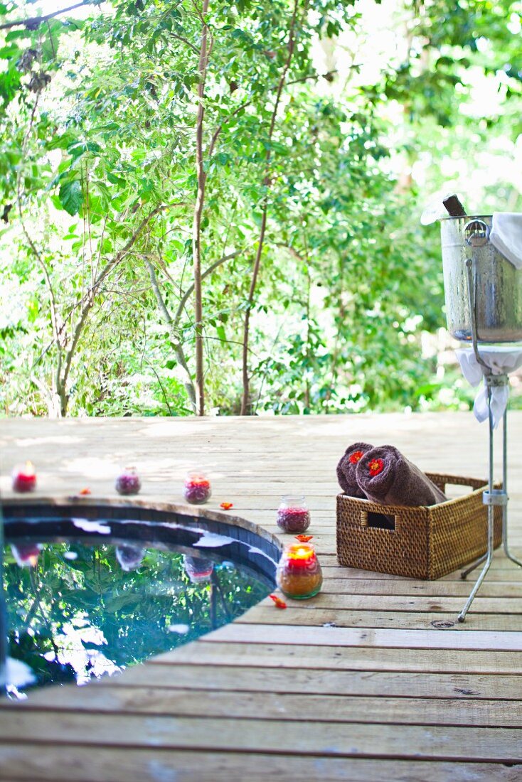 Wooden terrace with round pool, basket of rolled towels and champagne bucket against backdrop of green trees