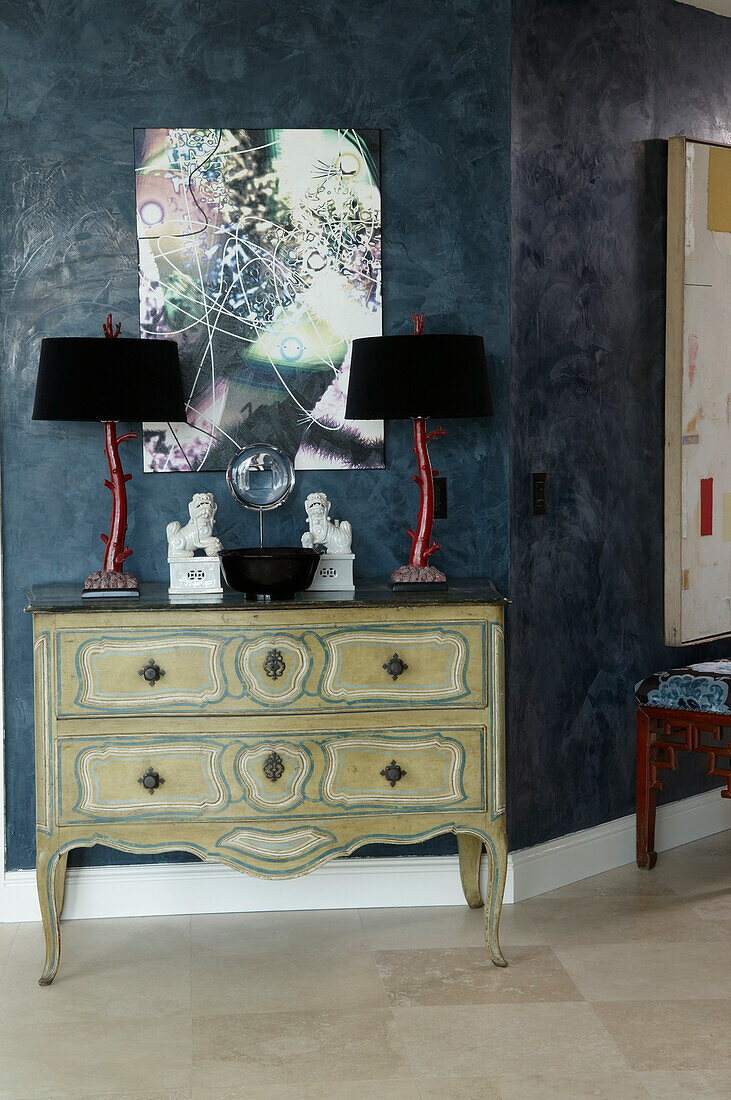 Painted chest of drawers on legs and two table lamps below abstract artwork on blue wall