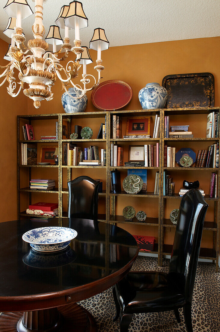 Round wooden table with heavy base and chairs with patent leather covers on leopard-print rug in front of shelving against ochre yellow wall