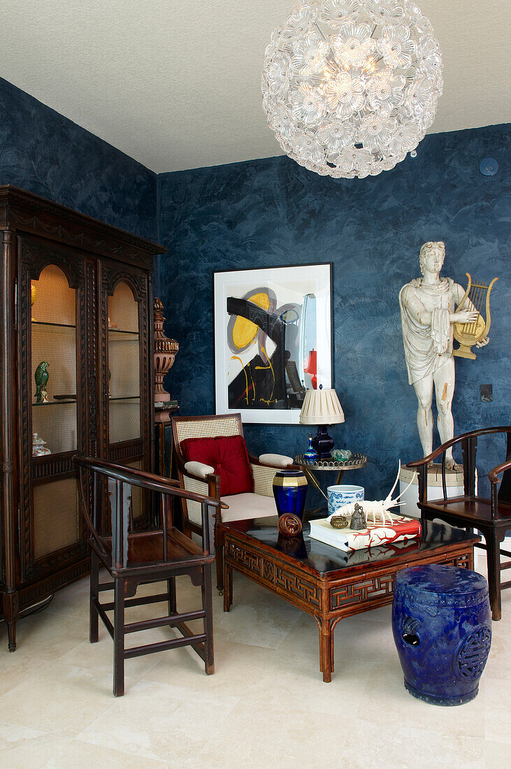 Modern spherical pendant lamp above Oriental coffee table and dark wooden chairs in front of stone sculpture in corner of blue living room