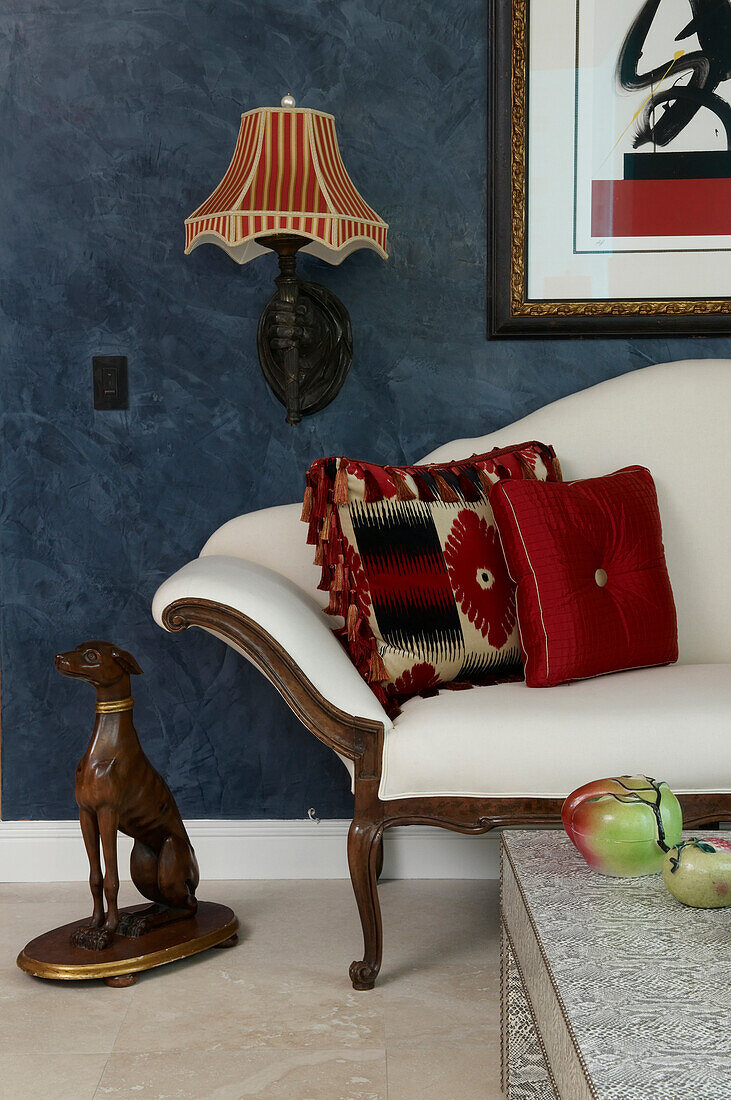 Detail of upholstered bench with scatter cushions leaning on arm next to animal figurine against blue marbled wall with traditional sconce lamp