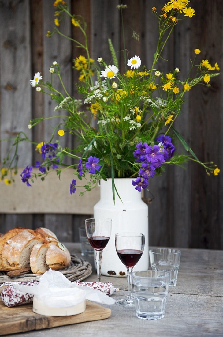 Käse, Brot, Rotwein und Milchkanne mit Wiesenblumen auf rustikalem Holztisch