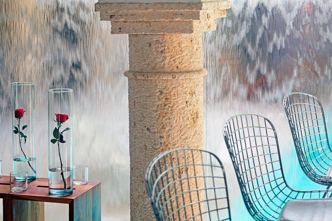 Wire chairs, roses in glass vases on side tables and stone columns in front of wall with waterfall effect