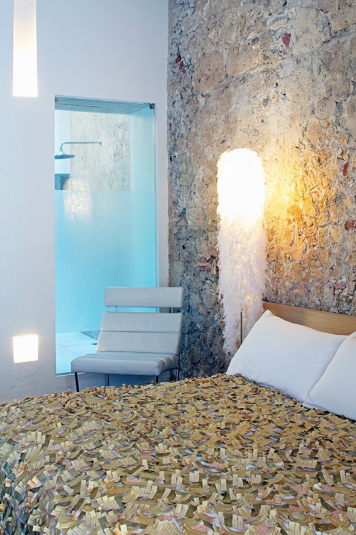 Doppelbett mit Designer-Tagesdecke vor Natursteinwand und moderner, gepolsterter Stuhl vor Innenfenster mit Blick in Duschbereich
