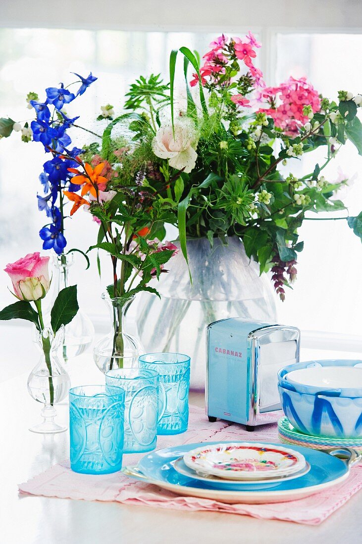 Blaue Gläser, Serviettenspender und bemaltes Keramikgeschirr vor Vasen mit Sommerblumen