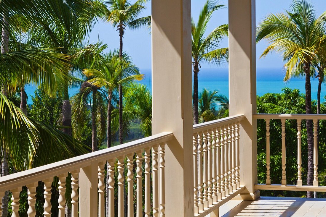 Veranda with white, carved wooden balustrade and sea view through palm trees
