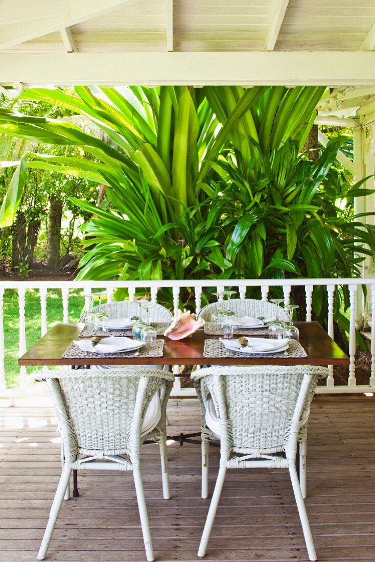 weiße Rattanstühle um Tisch mit Gedecken auf Holzboden einer Veranda vor tropischem Palmenbusch