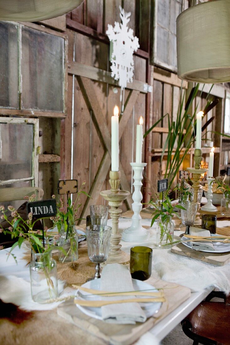Festlich gedeckter Tisch mit brennenden Kerzen und Wiesenblumen in Gläsern vor rustikaler Holzwand