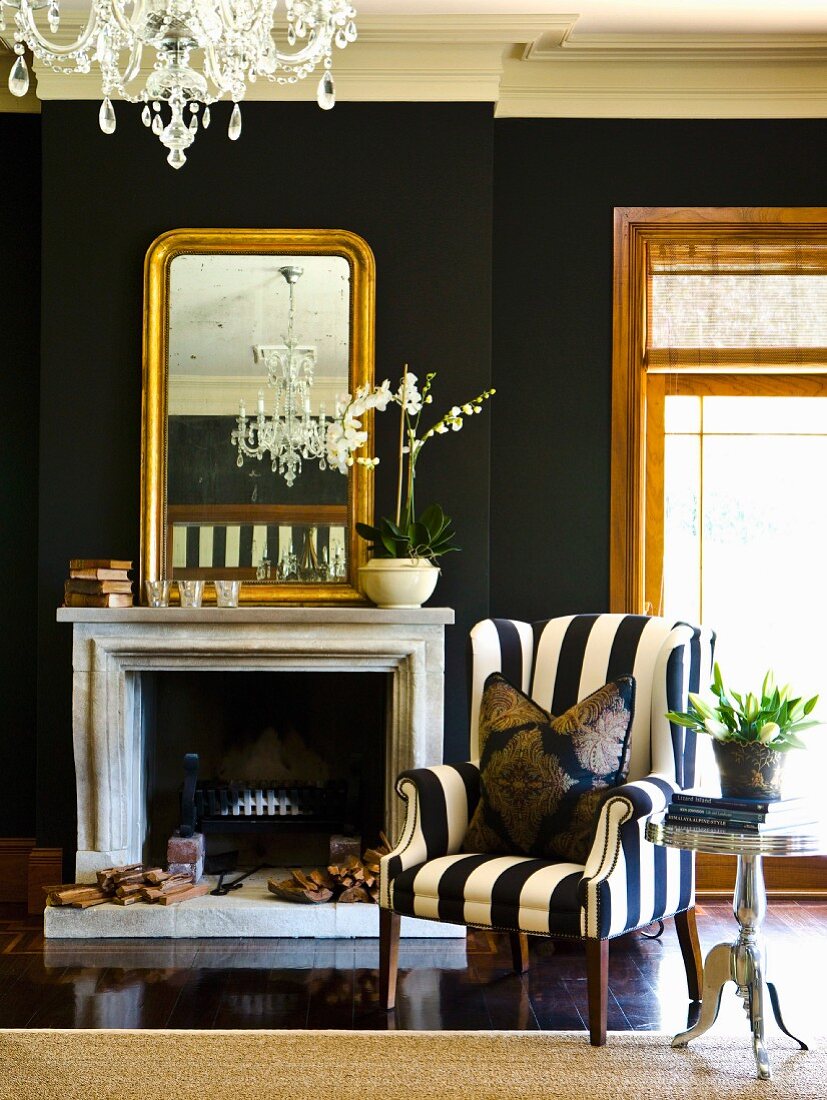 Schwarz-weiss gestreifter Sessel neben offenem Kamin und Spiegel mit Goldrahmen an schwarz getönter Wand