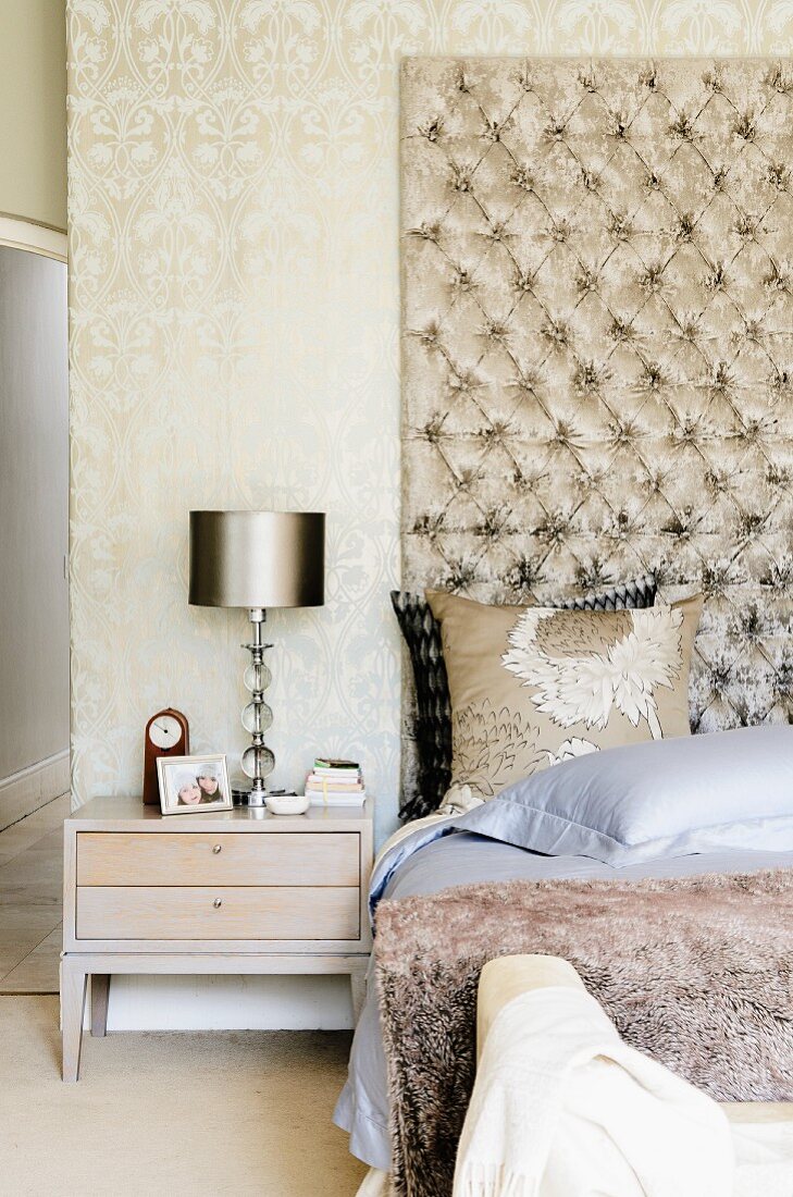 Herrenschlafzimmer in hellen Brauntönen - Bett vor hohem gepolstertem Wandpaneel und Fell Tagesdecke neben Nachtkästchen mit Tischleuchte