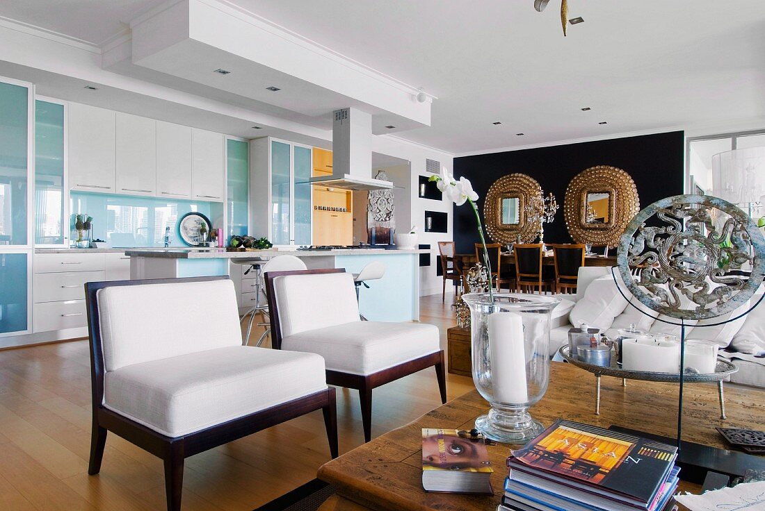 Eclectic mixture of styles in open-plan interior with classic upholstered chairs; kitchen with island counter and gilt-framed mirrors on black wall in background
