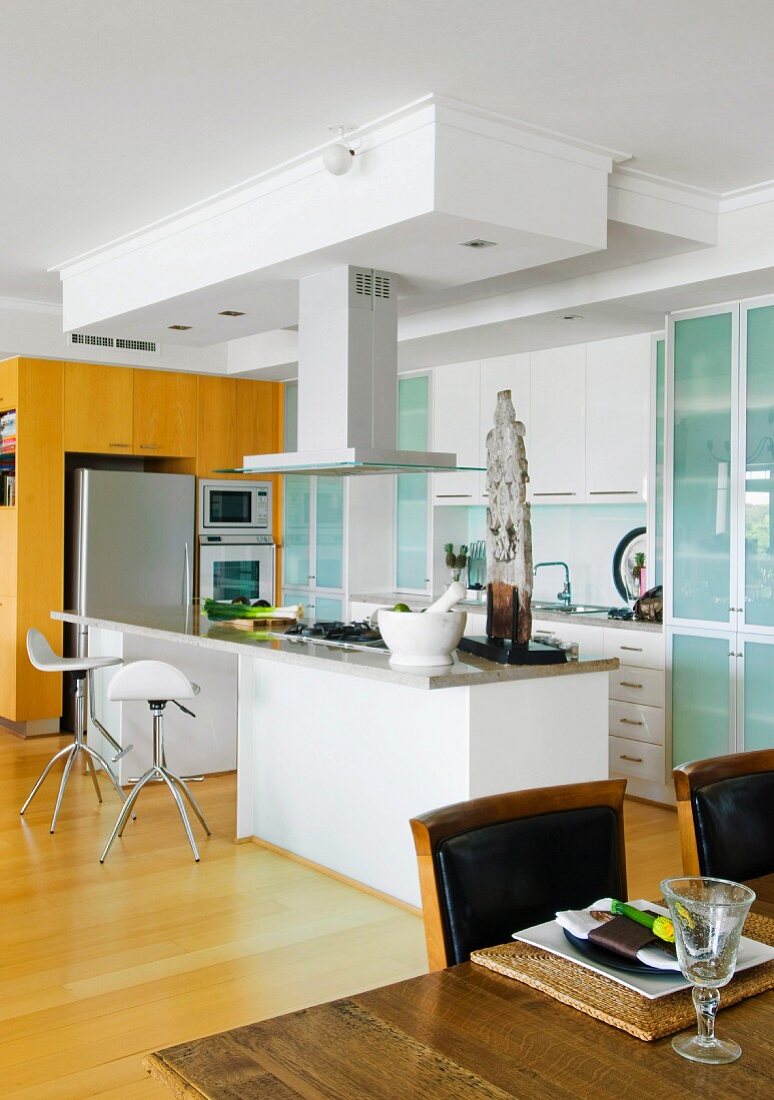 weiße Einbauküche mit grün schimmernden Glastüren und Küchentheke; im Vordergrund Esstisch mit Gedeck