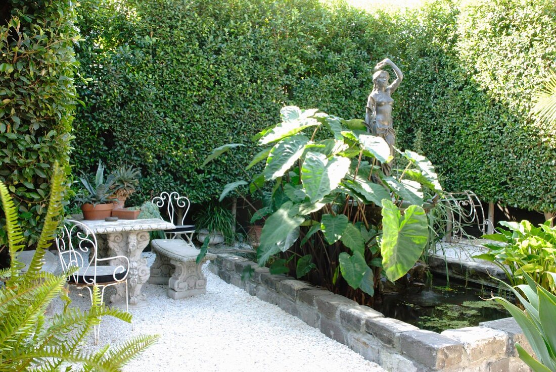 Small courtyard with lush greenery, pond, statue of woman and collection of antique-style garden furniture