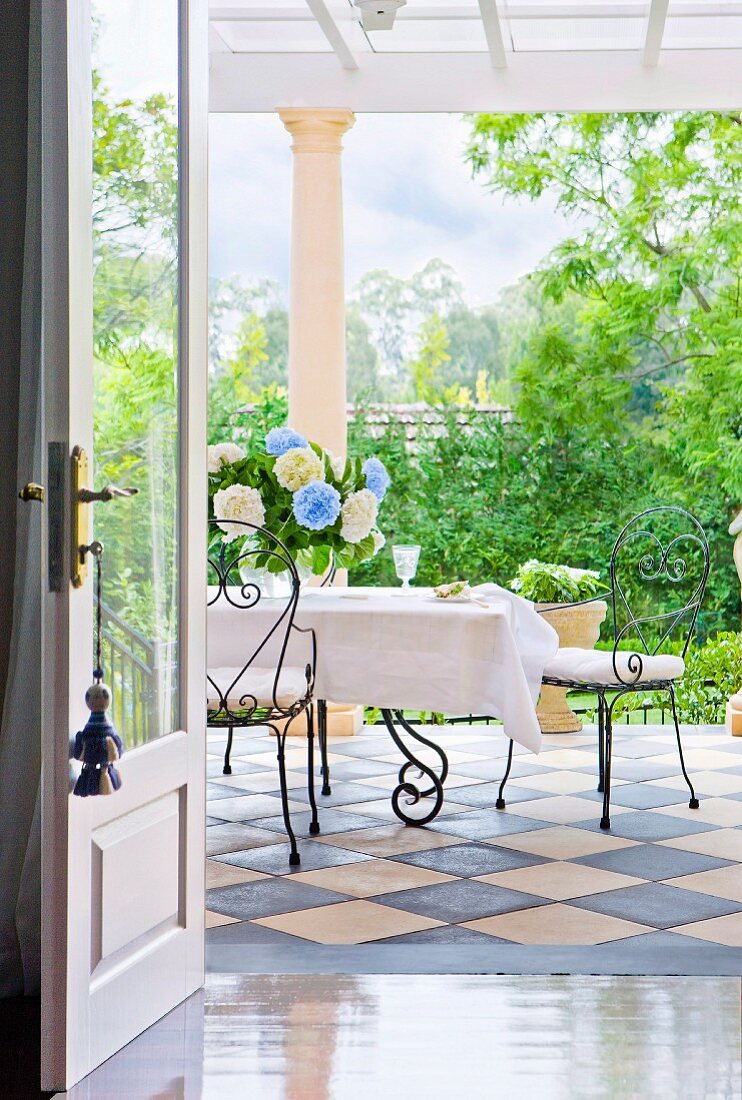 Blick durch offene Tür auf Terrasse mit Schachbrettfliesen und filigranen Drahtstühlen an weiss gedecktem Tisch