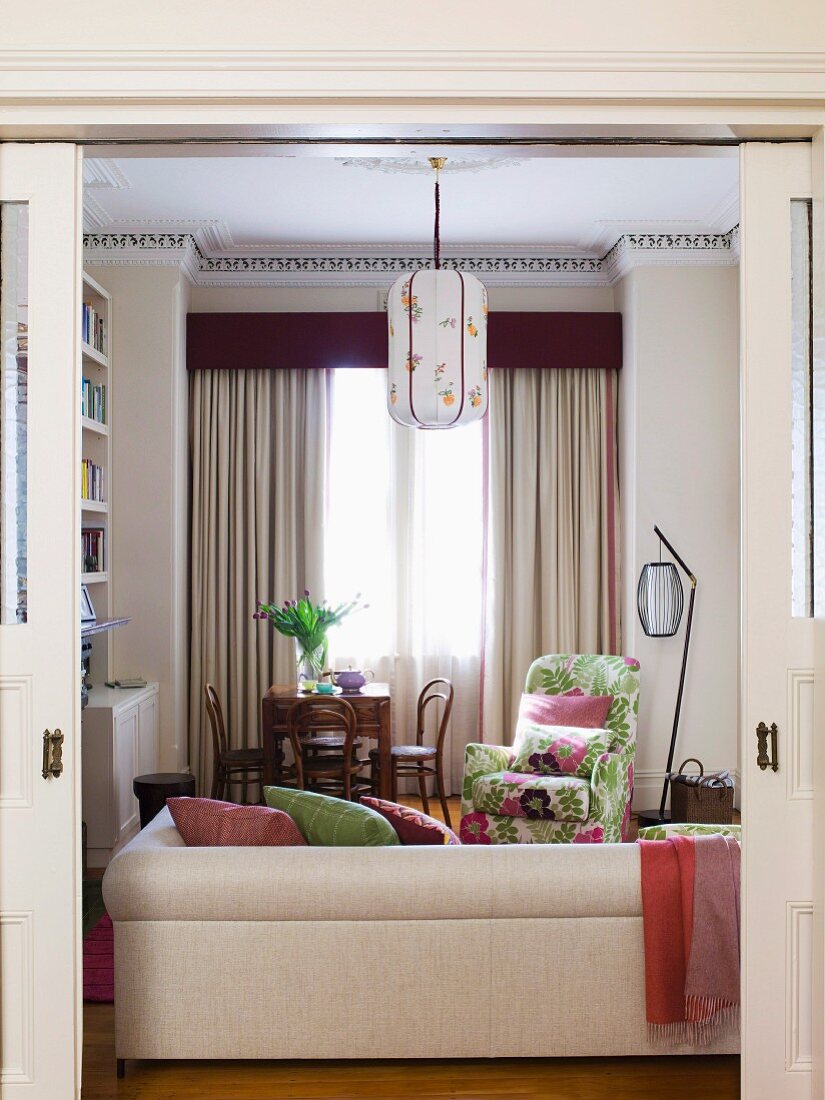 Blick durch offene Schiebetür auf helle Polstercouch und geblümten Sessel in Loungebereich vor Fenster mit bodenlangem Vorhang