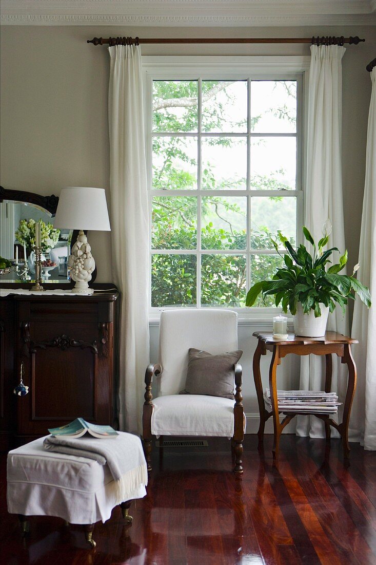 Traditional sitting area by a window with an armchair between a chest of drawers with a mirror and a side table