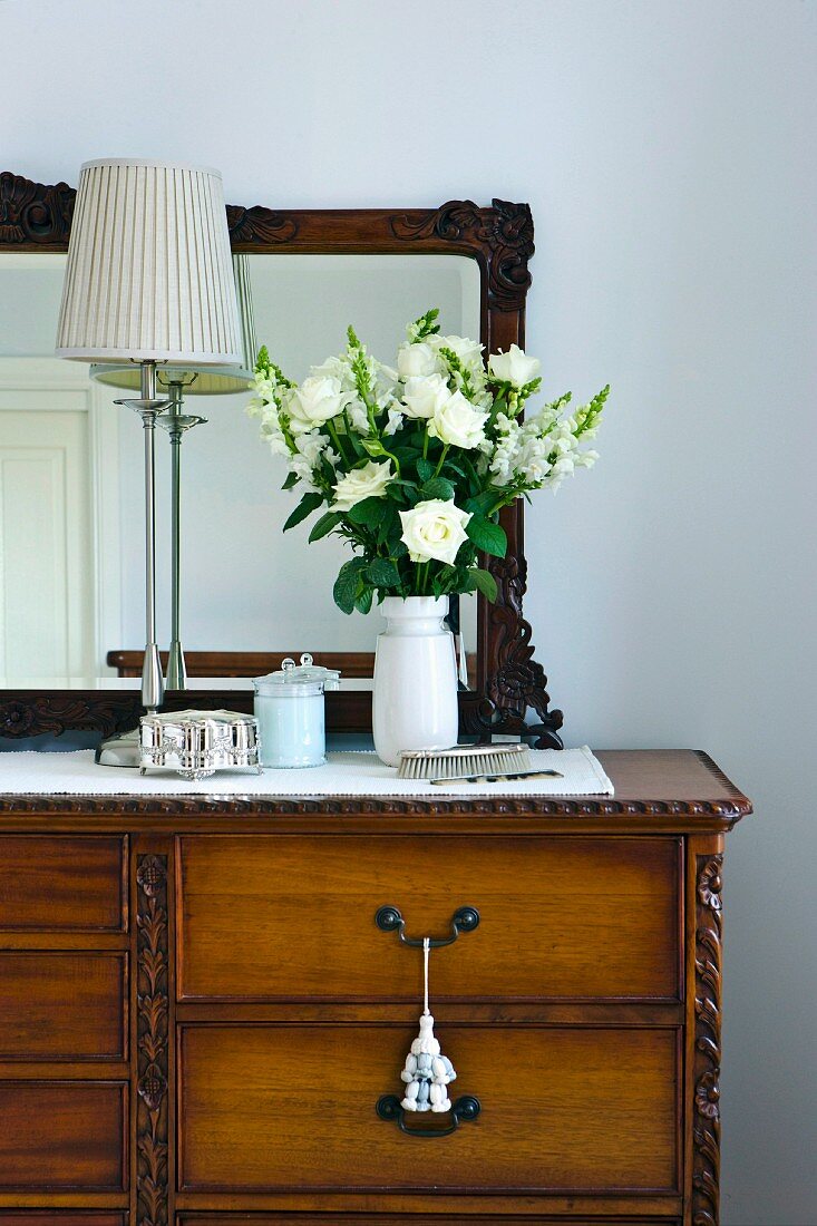 Bouquet of white flowers and table lamp on an antique chest of drawers with mirror