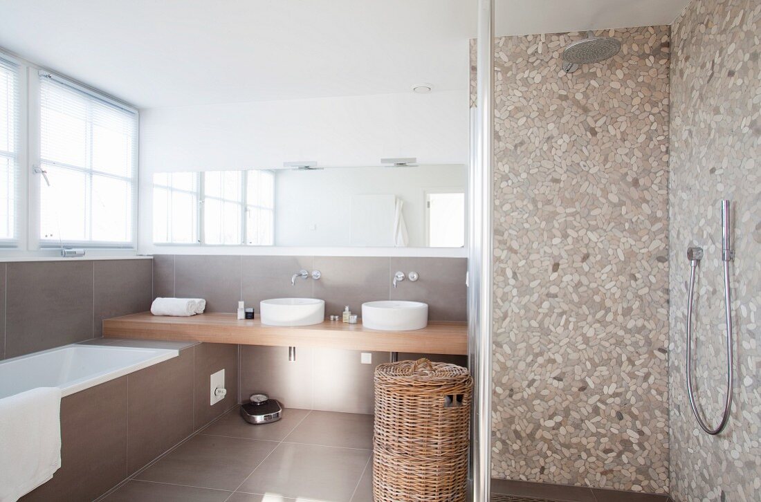 Shower with terrazzo-style tiles and modern washstand in background with two sinks below wide mirror on wall