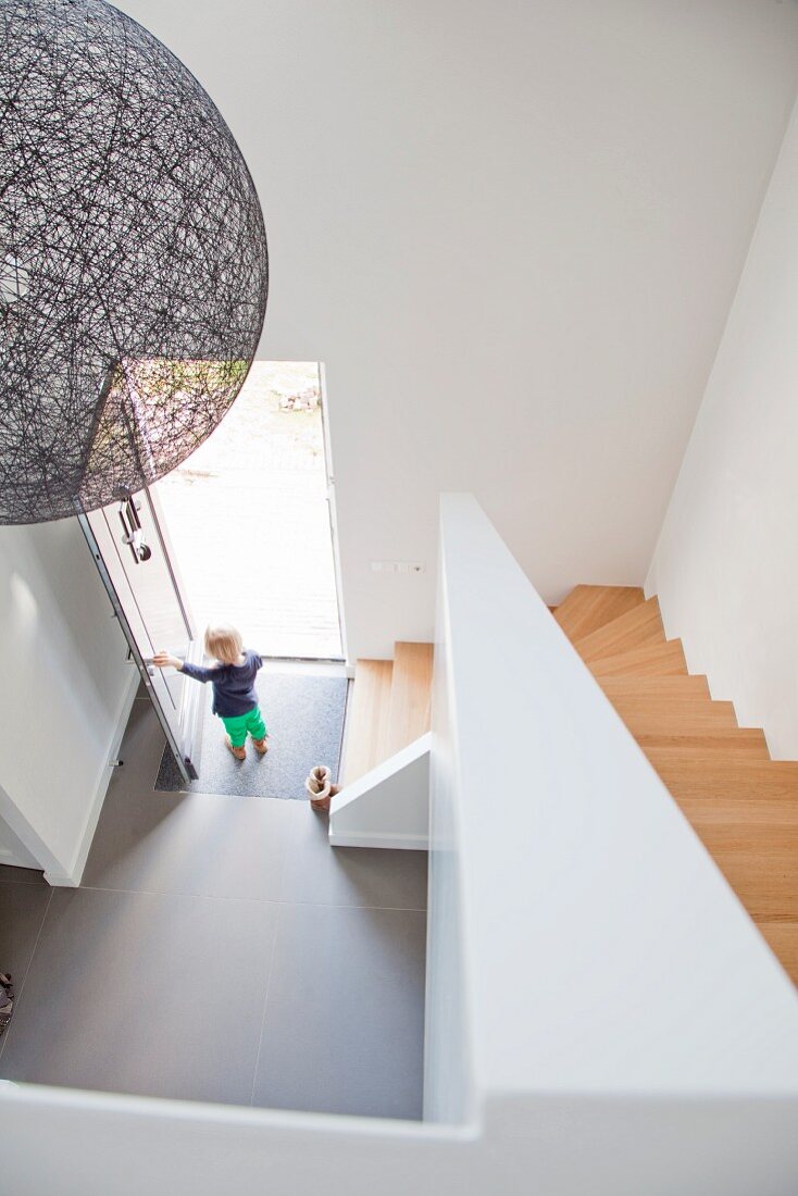 Dramatischer Blick von oben auf Designerhängeleuchte neben Treppenabgang und kleines Kind steht in offener Haustür