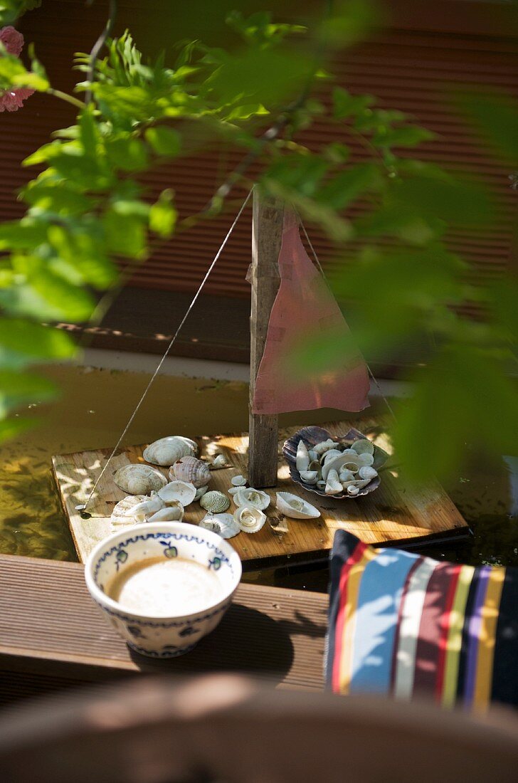 Schale neben Kissen auf Holzbank und selbstgebautes Miniatur Segelboot im Wasserbecken