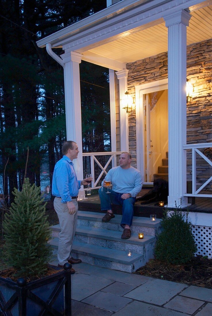 Two men drink the front of the lighted porch of a house