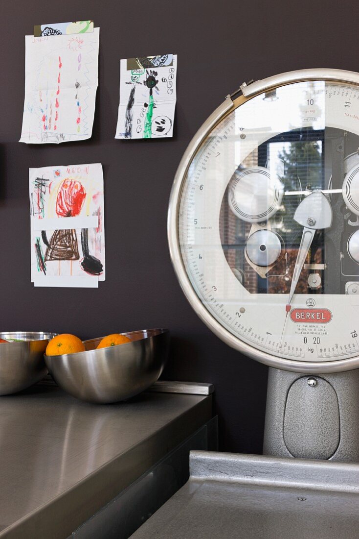 Old industrial scales next to bowl of oranges on stainless steel worksurface and children’s drawings