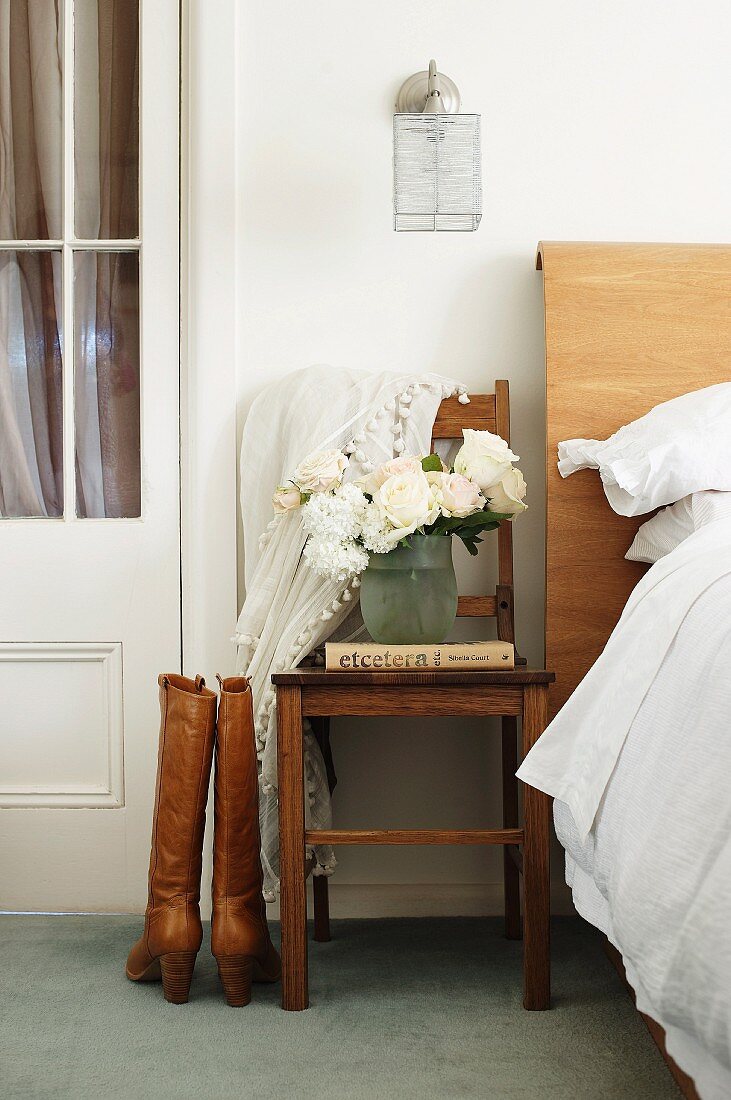 Braune Damenstiefel neben Stuhl mit Blumenvase und teilweise sichtbares Bett mit Kopfteil aus Holz