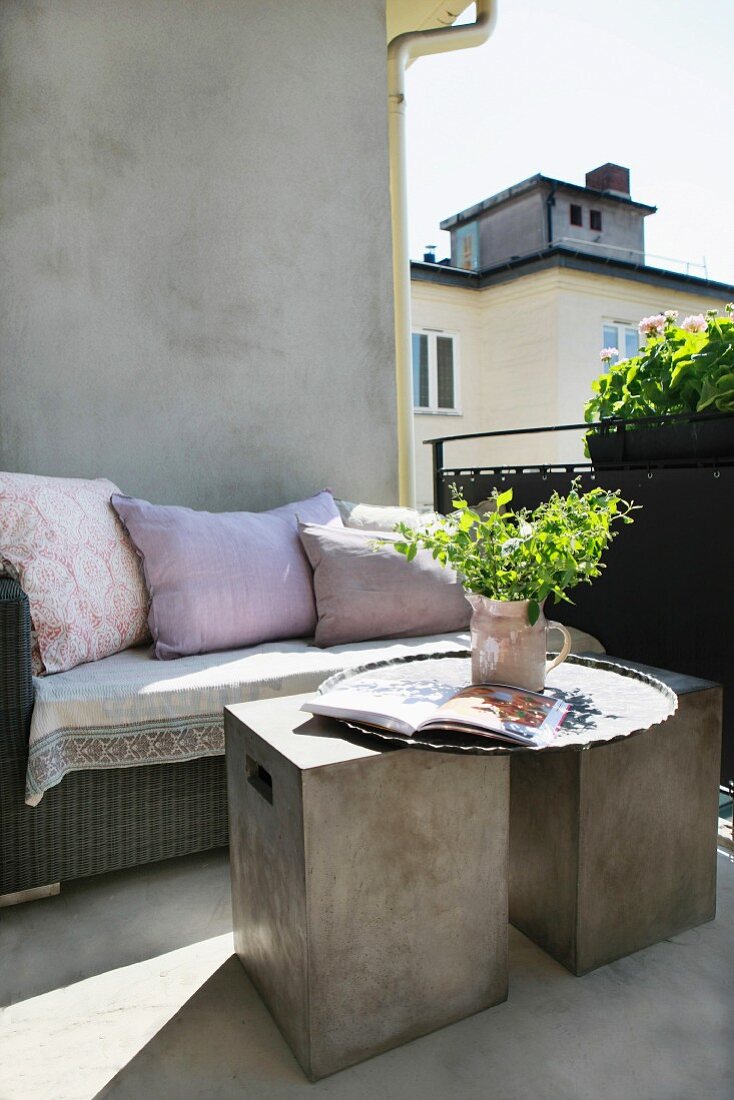 Sunny balcony with modern concrete cubes as side tables in front of rattan bench with cushions against house facade