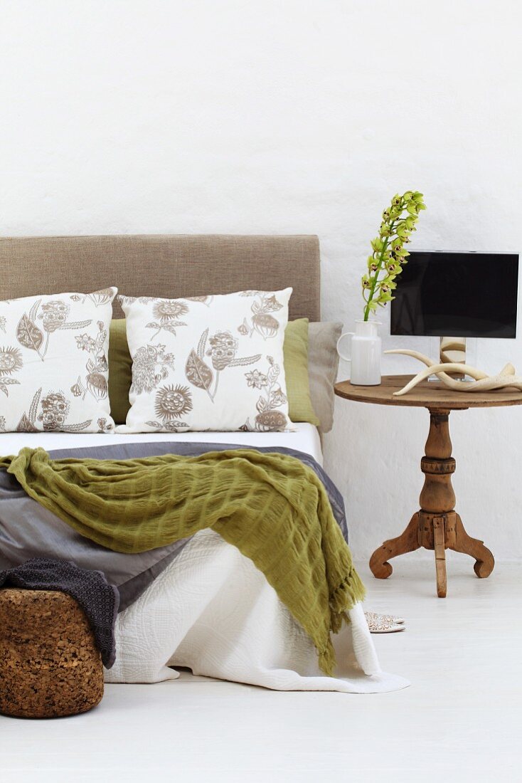 Tagesdecken und Kissen auf modernem Doppelbett mit Kopfteil neben rustikalem Beistelltisch aus Holz mit gedrechseltem Fuss