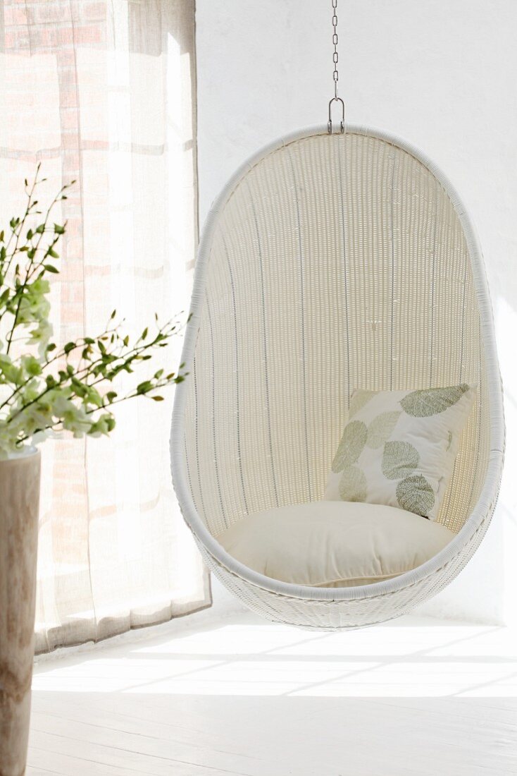 Gemütlicher Hängestuhl aus weißem Geflecht in Zimmerecke neben Fenster mit geschlossenem, transparentem Vorhang und Lichtspiel auf Boden
