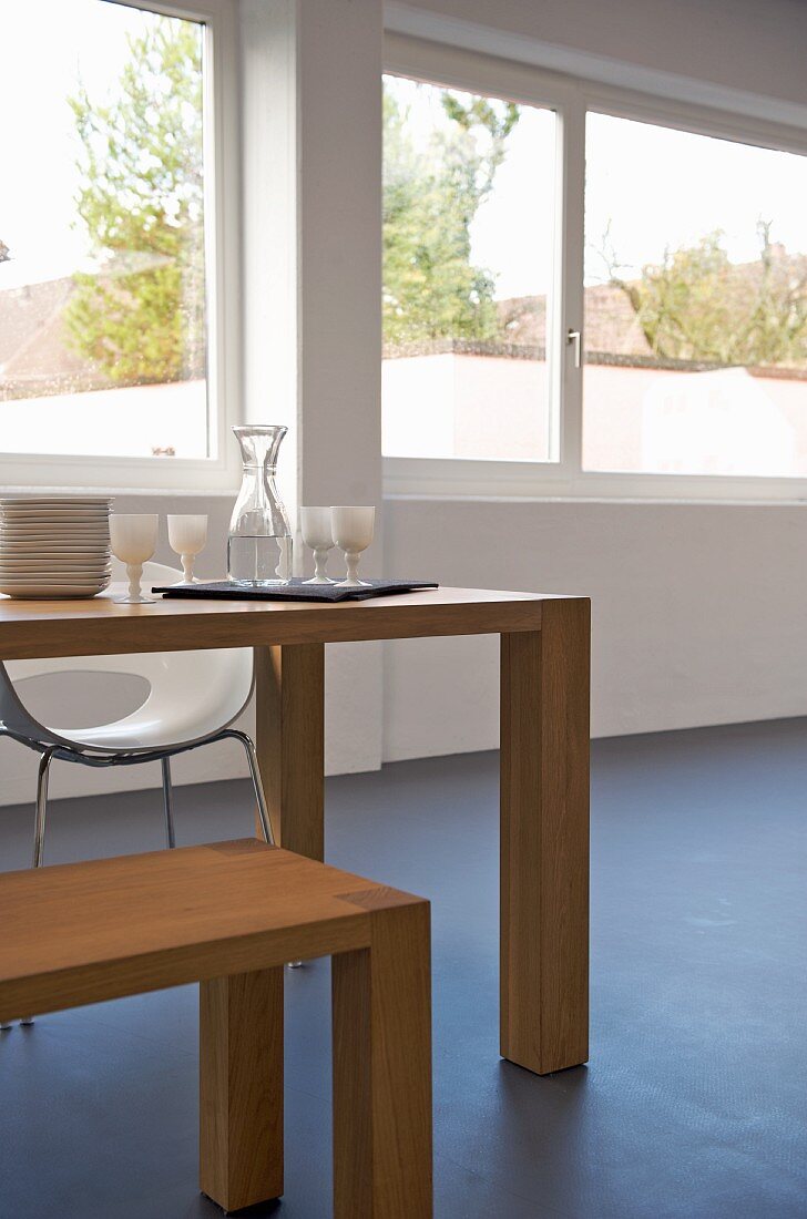 Crockery and glasses on minimalist dining table with matching wooden bench in front of bank of windows