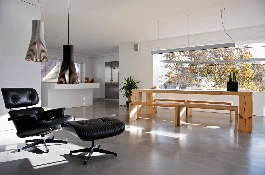 Klassiker Sessel mit schwarzem Lederbezug und passendem Fussschemel vor moderner Esstischgarnitur aus Holz in offenem Wohnraum