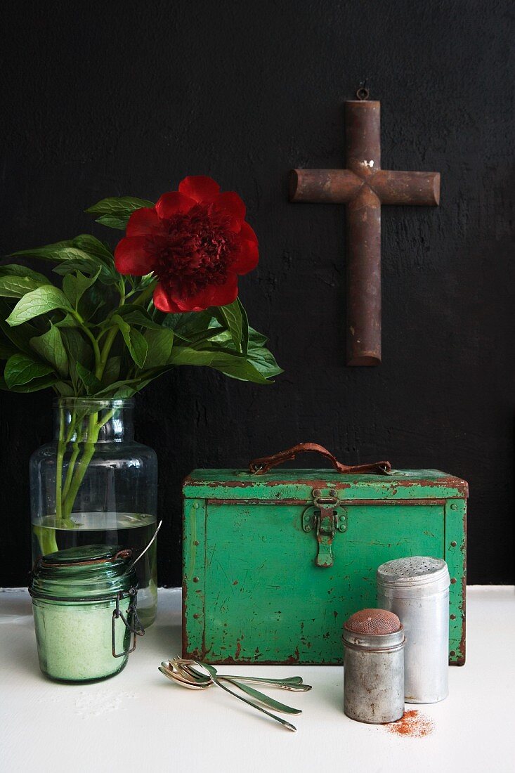Vintage Küchenutensilien vor grünem Metallkoffer und Vase mit roter Blume vor schwarzer Wand mit aufghängtem Kreuz