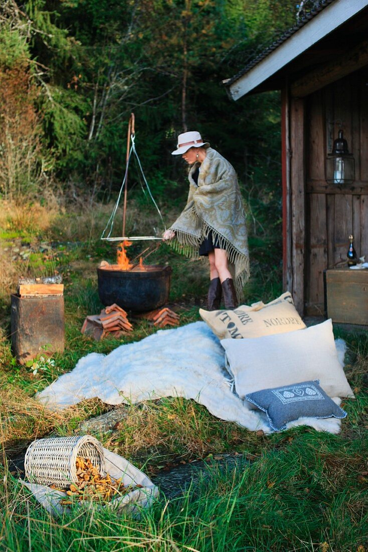 Lager mit Kissen und Felldecke vor einem Unterstand im Wald; Frau mit Hut und umgehängter Decke an einem Feuertopf