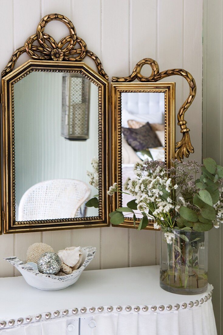 Blumenstrauss in Glasvase auf geschwungenem Wandtisch mit weißem Bezug und Ziernieten vor mehrteiligem Spiegel mit Goldrahmen an Holzwand