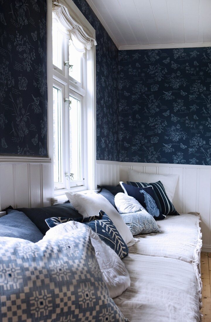 Kissen mit blau-weißem Muster und Polster auf Sitzbank am Fenster und blaue, gemusterte Tapete an Wand