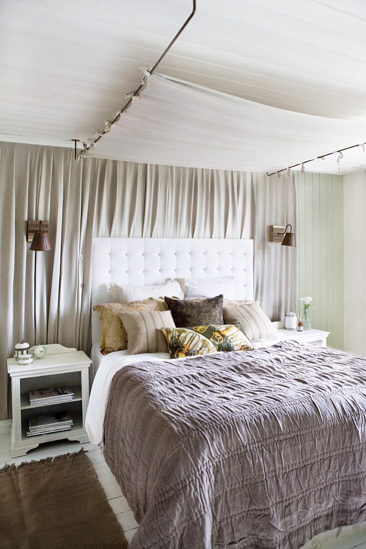 Graue Tagesdecke auf Doppelbett mit gepolstertem Kopfteil vor hellgrauem Vorhang an Wand und Lichtsegel unter der Decke