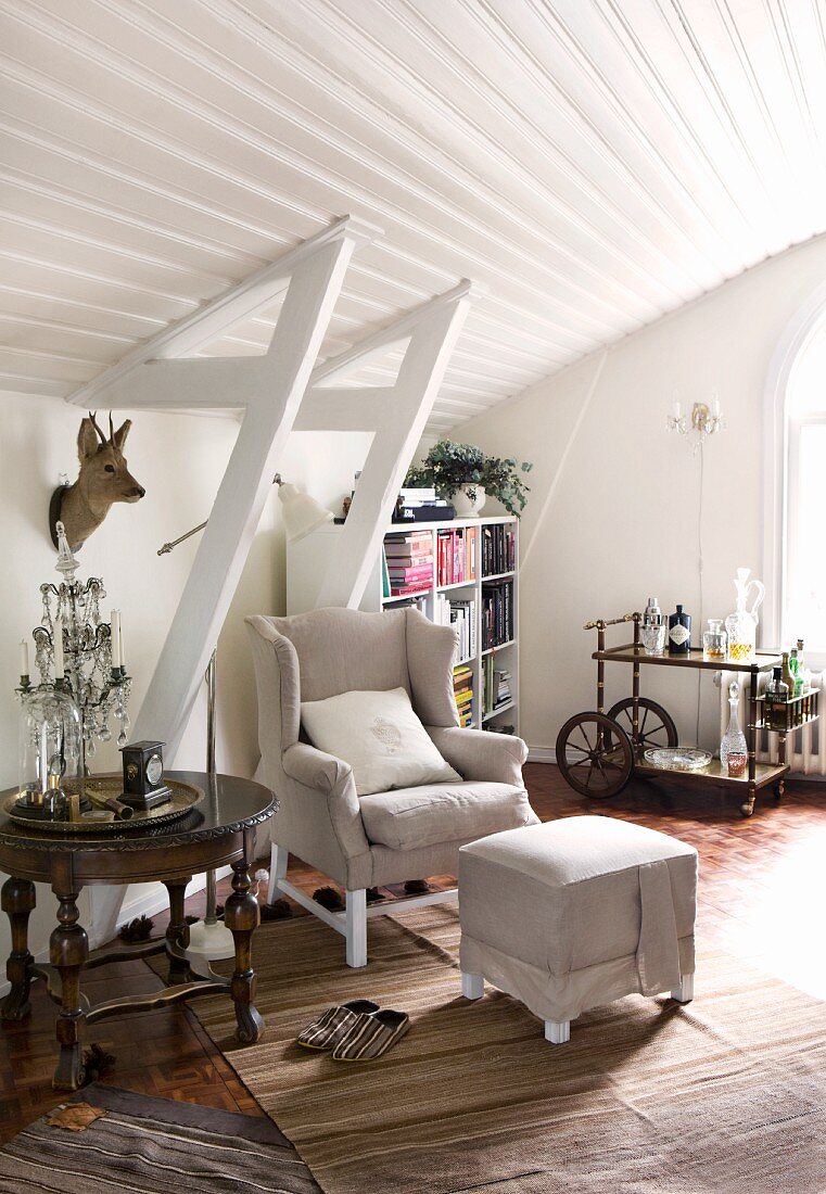 Wing-back chair and footstool, antique table and stuffed deer's head in white, wood-clad attic interior