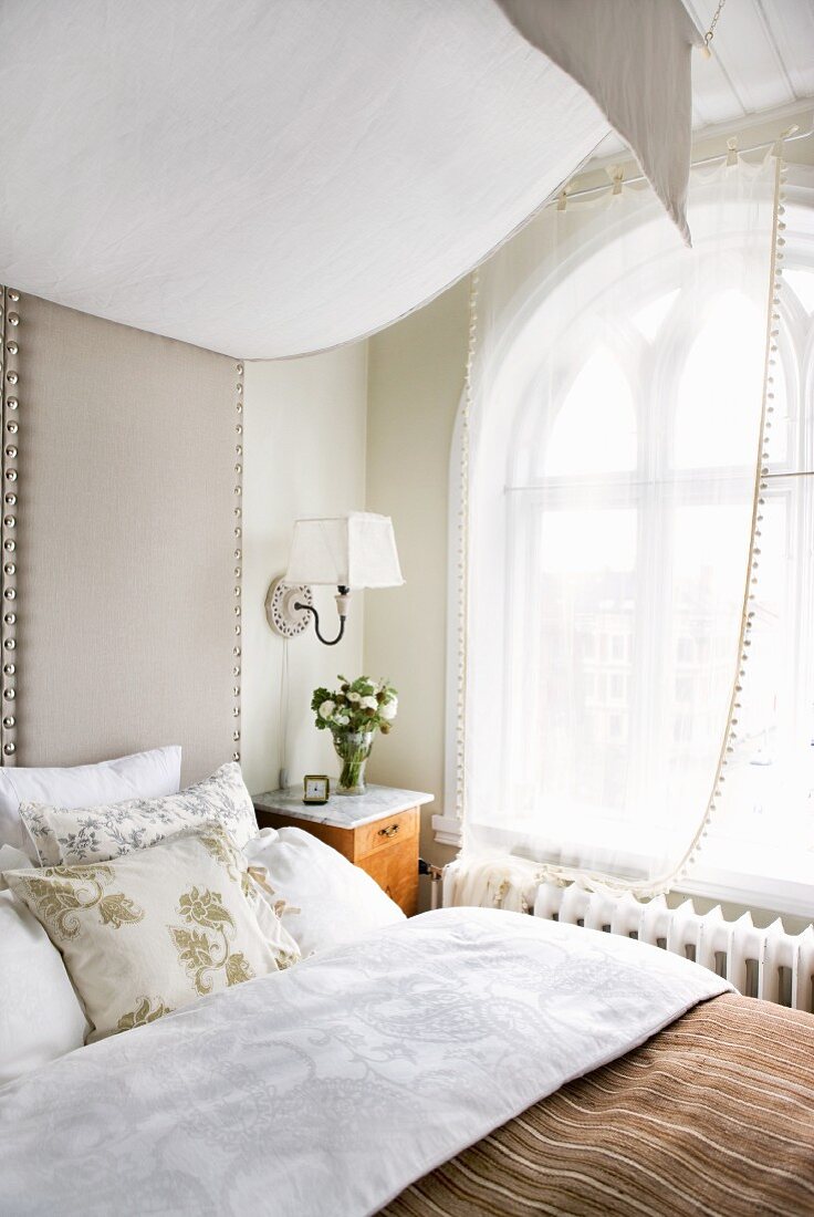 Bett mit gepolstertem Kopfteil und Stoffbaldachin vor Rundbogenfenster