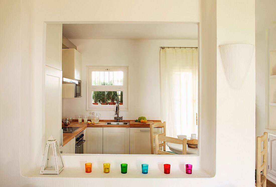 Colorful glass candle holders on a shelf in front of an wide serving hatch and view into a bright, modern kitchen