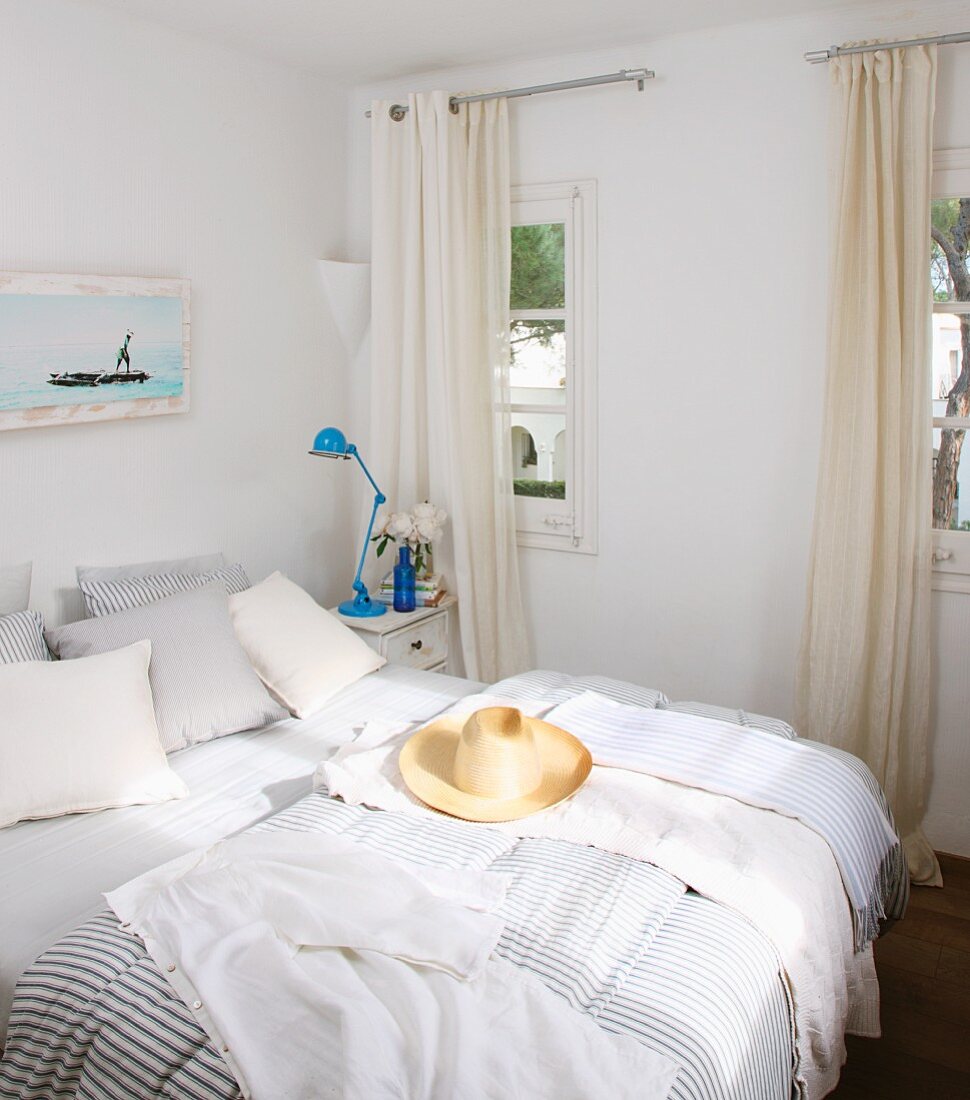 Strohhut auf Doppelbett mit gestreifter Bettwäsche neben Fenster mit bodenlangem Vorhang in weißem Schlafzimmer