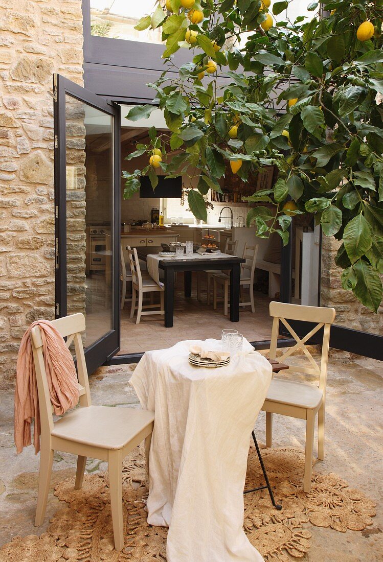 Hellgraue Küchenstühle und Tisch mit Tischtuch auf Terrasse unter Zitronenbaum vor offenen Terrassentüren und Blick auf Essplatz