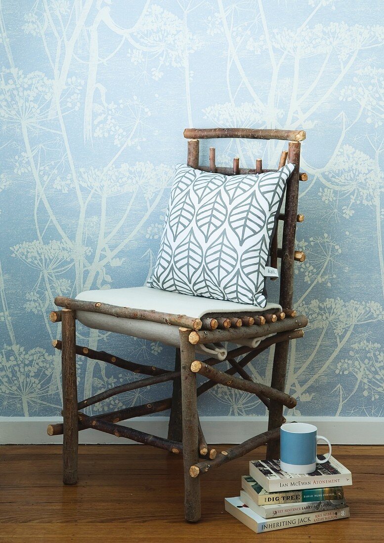 Kissen auf selbstgebautem Stuhl aus naturbelassenen Ästen neben Bücherstapel auf Boden vor Wand mit weissen Blumenmotiven auf hellblauer Tapete