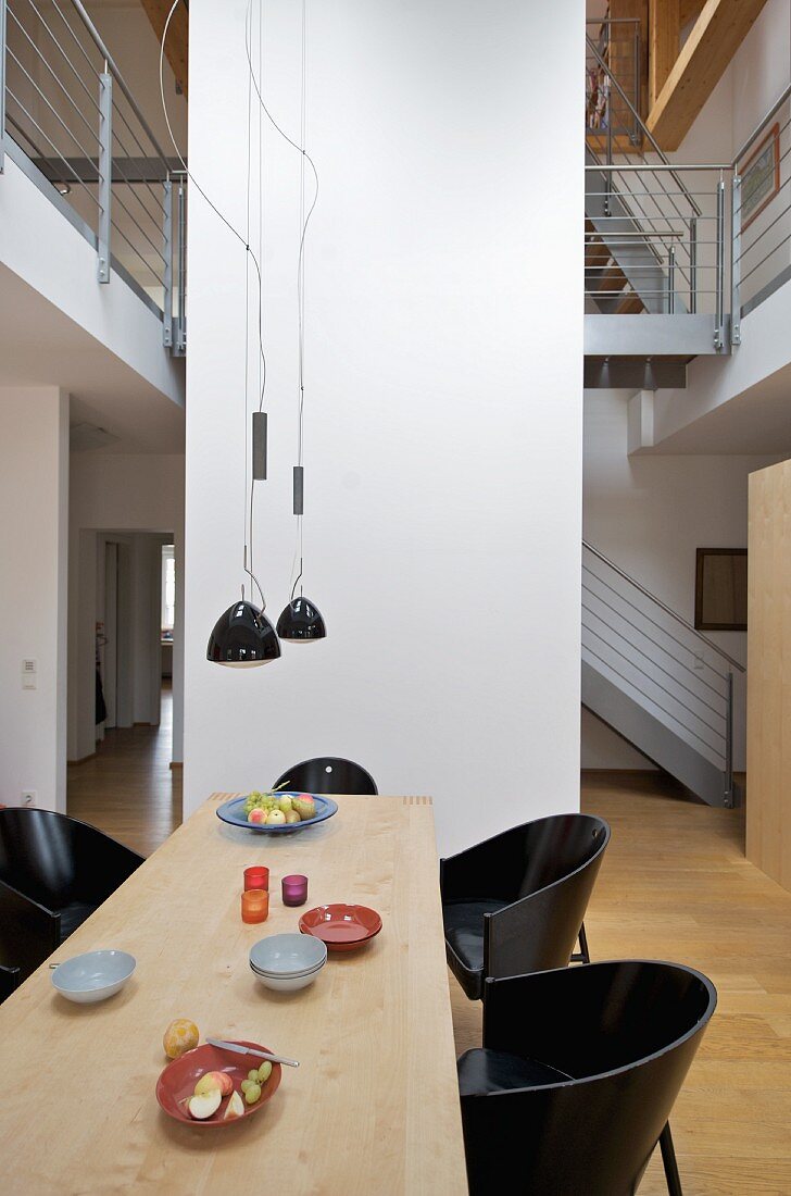Schalenstühle aus dunklem Holz an langem Esstisch unter zeitgenössischen Hängelampen vor Raumteiler in offenem Wohnraum und Blick auf Galerie
