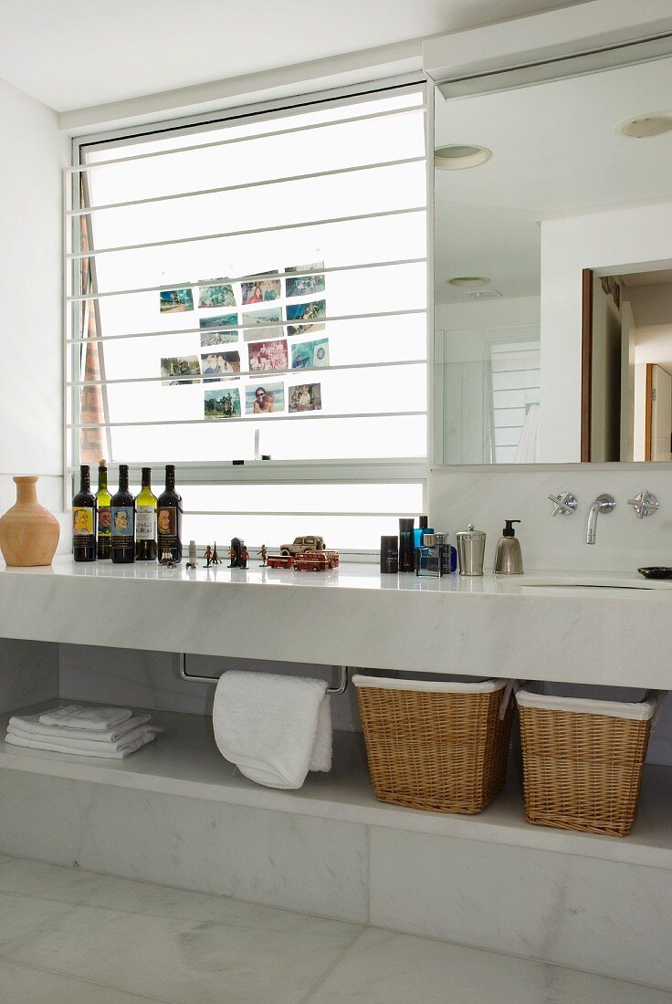 White marble bathroom with bottles on long washstand and photos on frosted glass window next to large wall-mounted mirror
