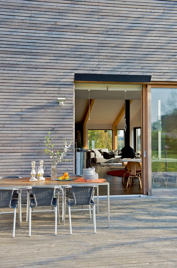 Tisch und Stühle auf Terrasse vor zeitgenössischem Haus mit Holzverschalung und Blick durch offene Terrassentür ins Wohnzimmer