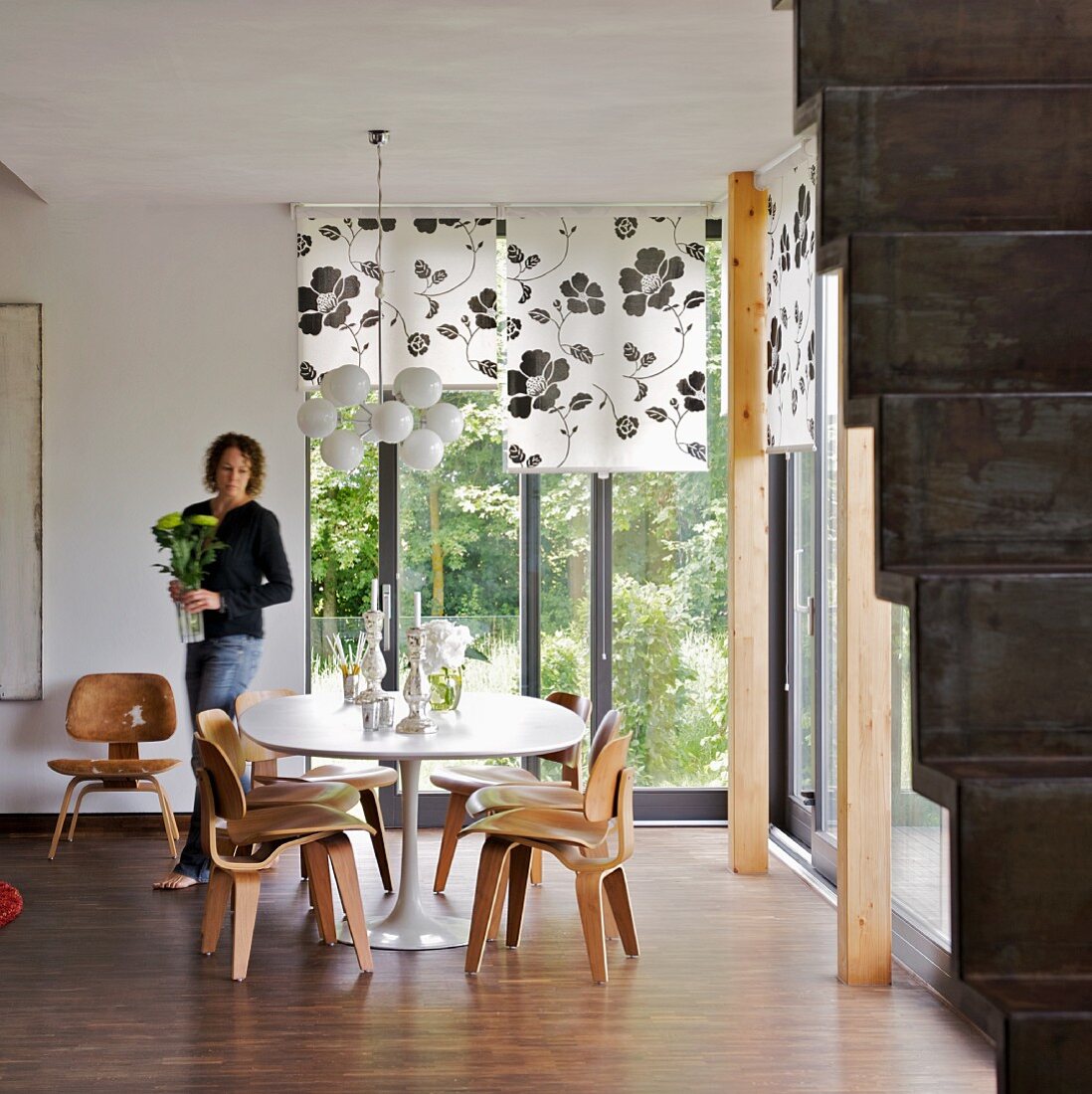 Sitzplatz mit Holzstühlen im 50er Jahre Stil in Zimmerecke mit Gartenblick und eine Frau mit Blumenvase in den Händen
