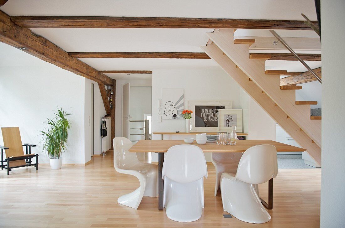Dining area with white plastic shell chairs below stairs in open-plan interior with visible wood structures on walls and ceiling